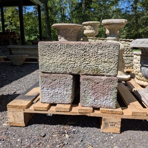 May include: A collection of weathered stone planters and rectangular blocks. The blocks have a rough, textured surface with shades of gray, green, and red. The items are displayed on a wooden pallet, suggesting outdoor use. Other decorative stone planters are visible in the background.