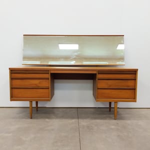 May include: A mid-century modern wooden desk with a rectangular mirror. The desk has three drawers on each side and is supported by four tapered legs. The wood has a warm, natural finish, and the mirror is rectangular with a wooden frame.