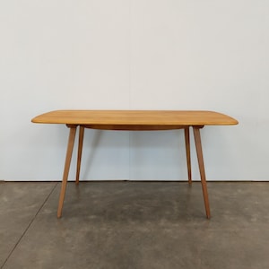 May include: A light brown wooden table with a rectangular top and four tapered legs. The table has a mid-century modern design and is set against a white wall and a concrete floor.