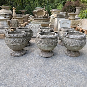 May include: A collection of weathered, gray stone garden planters featuring a rounded bowl shape, a pedestal base, and decorative scrollwork patterns around the exterior. These heavy, textured concrete vessels are arranged in an outdoor setting.