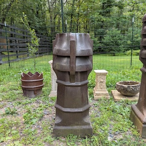 May include: A tall, dark brown, cylindrical chimney pot with a flared top and vertical structural ribs. The object features a wide base and a segmented body design, standing upright on a grassy surface with other garden ornaments visible nearby.