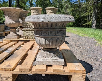 May include: A weathered, gray stone planter with a wide, decorative rim and a detailed base. The planter sits on a wooden pallet, with other similar planters visible in the background. The design features a Greek key pattern.