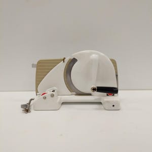 May include: A white and beige electric food slicer with a large circular blade. The slicer has a black handle and a red button. The base is white, and the blade guard is beige.
