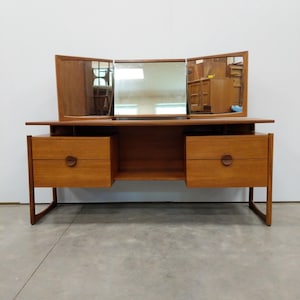 May include: A wooden vanity table featuring a light brown finish, a central rectangular mirror flanked by two adjustable side mirrors, and two sets of two drawers on either side. Each drawer is equipped with a circular wooden pull handle.