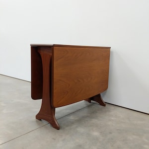 May include: A brown wooden drop-leaf table with a smooth rectangular top and two hinged side panels folded down. The table features a sturdy base with two curved, flared legs on each side, showcasing a natural wood grain finish throughout.