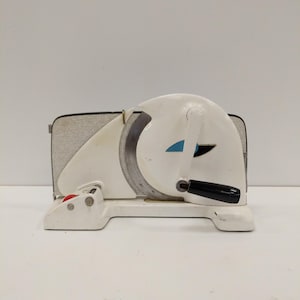 May include: A vintage white meat slicer with a black handle and a red button. The slicer has a circular blade and a textured side panel. A blue accent is visible on the white body.