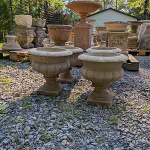 May include: A collection of weathered stone garden planters featuring a rounded bowl shape with a fluted base and a square pedestal. The planters are light gray and beige in color, showing signs of age and texture. Several similar stone vessels are arranged in the background.