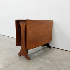 May include: A brown wooden drop-leaf table with a rectangular top and two hinged side panels folded down. The table features a smooth grain finish and is supported by two curved, flared legs that provide a stable base for the structure.