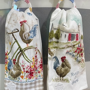 May include: Two kitchen towels with crocheted hangers. The towel on the left features a rooster riding a bicycle with a floral design. The towel on the right features a rooster with a barn in the background and the text "Rise & Shine". Both towels have a 2 in 1 functionality label.