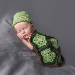 May include: A newborn baby wearing a green crocheted turtle costume with a brown and green shell pattern. The baby is sleeping on a gray blanket.