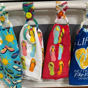 May include: Four colorful crocheted towel holders with a ring for hanging towels. Each holder features a different design with flip flops and summery themes. The towels have various designs including a sun, sunglasses, and the text "Better in Flip Flops".