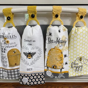 May include: Four yellow crocheted towel hangers with black buttons hanging on a white oven door. The towels have bee-themed designs with text that reads "Bee Rules", "Sweet as can", "Bee Happy", and "bee happy".