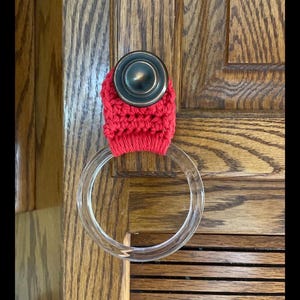 May include: A red crocheted door handle cover with a clear acrylic towel ring. The cover is wrapped around a bronze-colored door handle. The background is a wooden door with a natural wood grain.