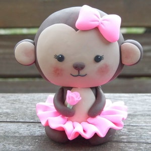 May include: A handcrafted figurine of a monkey with a brown body, a light brown face, and a pink bow. The monkey wears a pink tutu and holds a pink rose. The figurine has rosy cheeks and is designed in a cute, cartoon style.