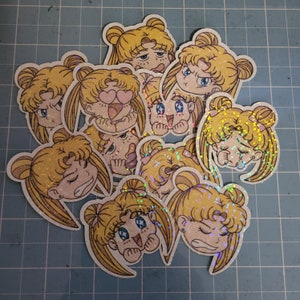 May include: A set of ten glitter stickers featuring different facial expressions of a cartoon character with blonde hair and pigtails. The stickers are printed on white paper and have a holographic finish.