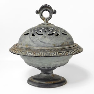 Puede incluir: Un quemador de incienso de hierro fundido gris con un diseño decorativo y una tapa abovedada con un asa. El quemador tiene una base con un patrón de clave griega.