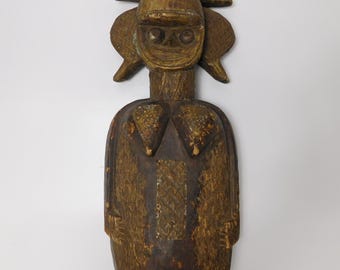 May include: A wooden sculpture of a stylized human figure with a crown-like headpiece. The figure is painted in shades of brown and gold, with intricate details carved into the surface.