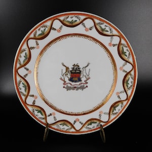 Könnte beinhalten: Ein weißer Porzellanteller mit goldfarbenem Rand und einem bunten Wappen in der Mitte. Der Teller hat einen dekorativen Rand aus goldenen und roten Blättern und Blumen.