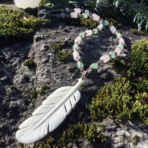 May include: A white carved feather pendant hangs from a necklace made of pink and green beads. The necklace is lying on a mossy rock.