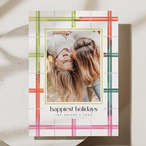 May include: Holiday card with a photo of two people, framed by a green border. The card has a white background with a plaid pattern in green, red, and pink. Text at the bottom reads "happiest holidays" and "The Smiths | 2024".