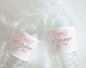 Puede incluir: Dos botellas de agua de plástico transparente con etiquetas rosas y blancas. Las etiquetas presentan un lazo rosa con el texto "Charlotte is One" en cursiva negra.