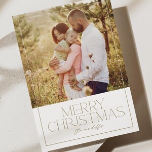 May include: A Christmas card with a photo of a family of three standing in a field. The card has the text "Merry Christmas" and "the maddolls" in a gold font.