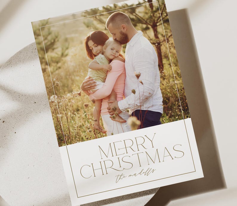 May include: A Christmas card with a photo of a family of three standing in a field. The card has the text "Merry Christmas" and "the maddolls" in a gold font.