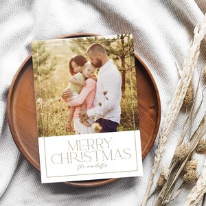 May include: A Christmas card with a photo of a family of three in a field. The card has a gold border and the text "Merry Christmas" in gold lettering. The family's last name is written in cursive below the greeting.