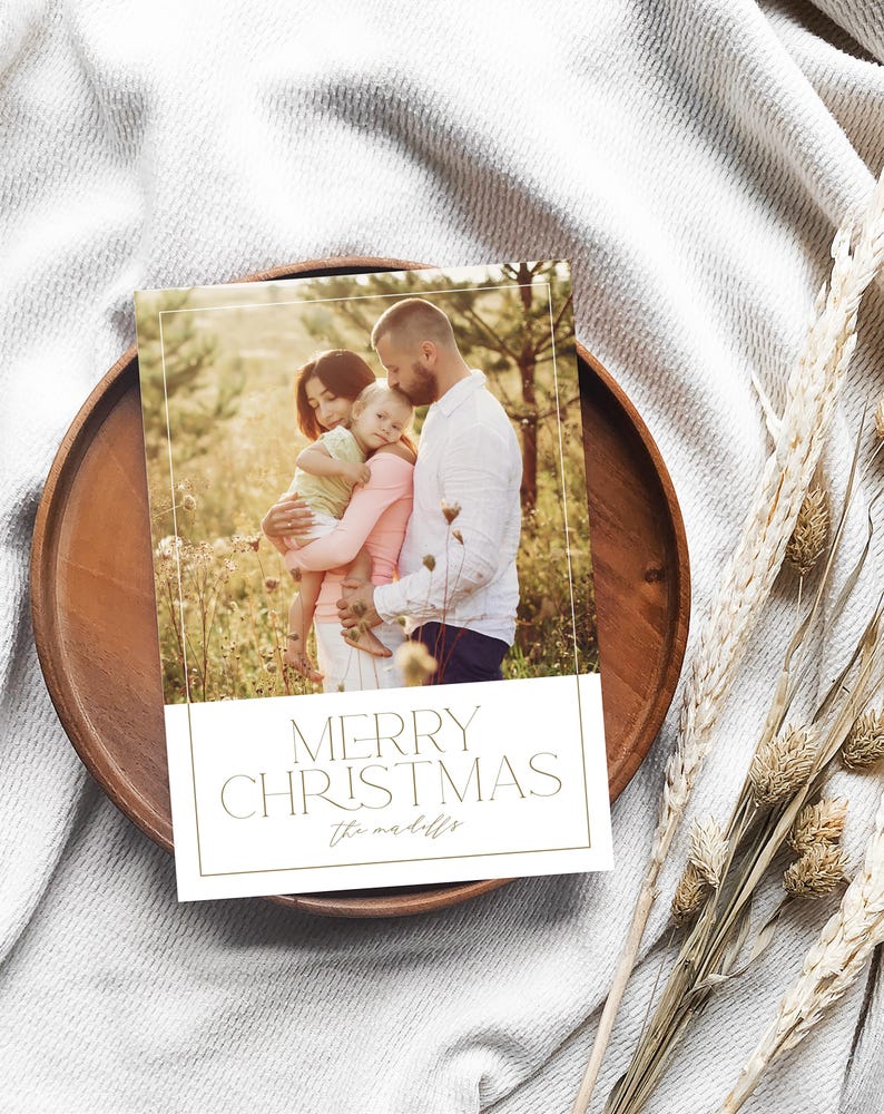 May include: A Christmas card with a photo of a family of three in a field. The card has a gold border and the text "Merry Christmas" in gold lettering. The family's last name is written in cursive below the greeting.
