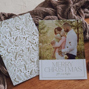 May include: A white Christmas card with a gold floral pattern on the left side and a photo of a family on the right side. The photo shows a family of three standing in a field. The text "Merry Christmas" is printed in a modern font on the card.