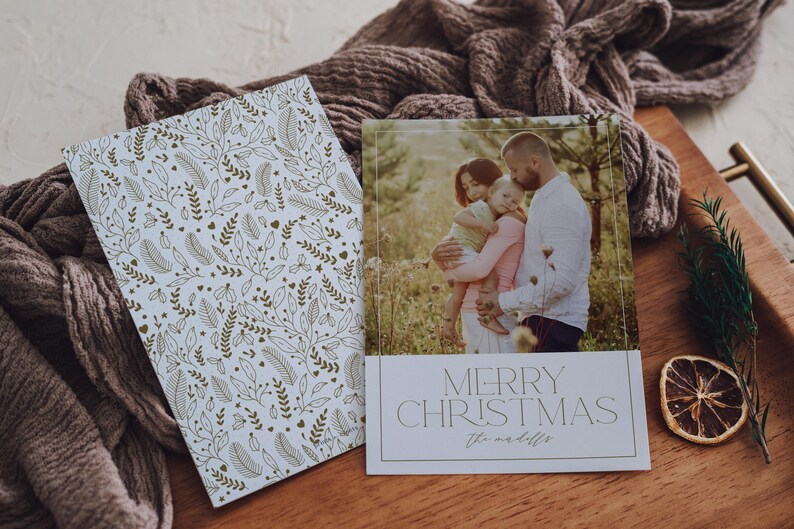 May include: A white Christmas card with a gold floral pattern on the left side and a photo of a family on the right side. The photo shows a family of three standing in a field. The text "Merry Christmas" is printed in a modern font on the card.