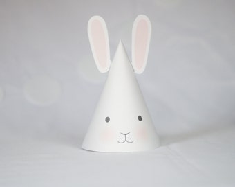 May include: White paper party hat shaped like a bunny with pink ears and black eyes. The bunny has a small gray nose and a pink mouth.