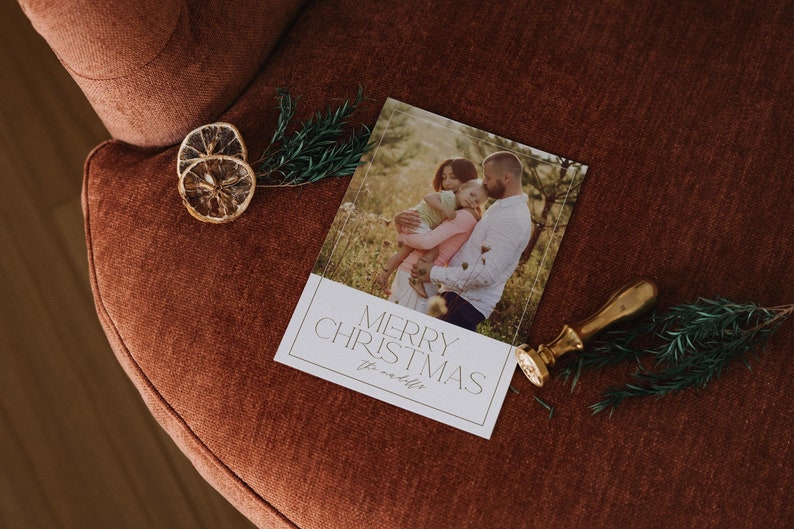 May include: A white Christmas card with a photo of a family of three in a field. The text "Merry Christmas" is printed in gold lettering on the card.