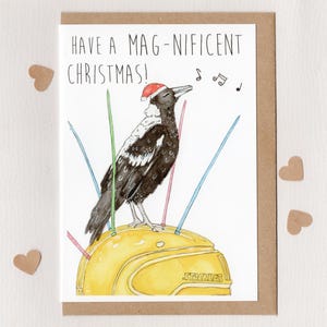 May include: Christmas card with a watercolor illustration of a black and white bird wearing a Santa hat, perched on a yellow object. The card reads "Have a Mag-nificent Christmas!" with musical notes.