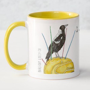 May include: White ceramic mug with a yellow handle and interior. The mug features a black and white bird illustration perched on a yellow object with colourful lines. The text "Barefoot Gilly Co" is printed on the side.