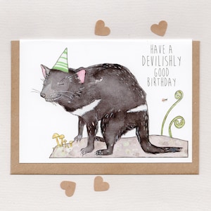 Puede incluir: Tarjeta de cumpleaños con una ilustración en acuarela de un demonio de Tasmania con un gorro de fiesta. La tarjeta tiene el texto "Have a devilishly good birthday". Pequeños recortes en forma de corazón marrón rodean la tarjeta.