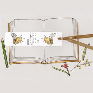 May include: A white bookmark featuring the text "BEE HAPPY" and two bee illustrations. A brown ribbon is attached to the bookmark. A green pencil and botanical drawings are also visible, set against an open book.