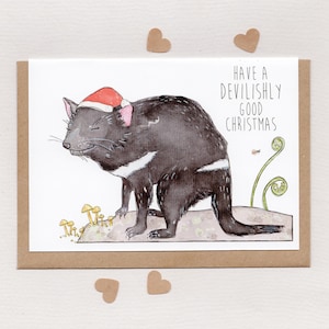 Puede incluir: Tarjeta de felicitación navideña con una ilustración en acuarela de un demonio de Tasmania con un gorro de Papá Noel. El texto dice "Have a devilishly good Christmas". La tarjeta está en un sobre marrón con adornos en forma de corazón.
