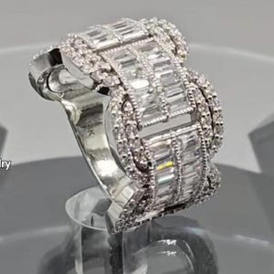 May include: A silver ring with a distinctive design, showcasing multiple rows of rectangular-cut stones. The ring is displayed on a clear stand, reflecting light. The brand name "VAMMJewelry" is visible at the top.