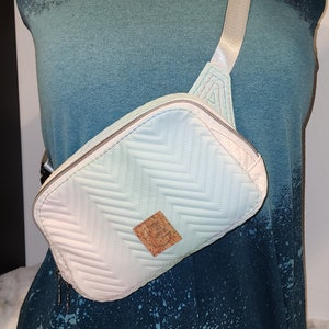 May include: A white quilted fanny pack with a chevron pattern and a cork patch with a crescent moon design. The fanny pack has a zipper closure and an adjustable strap.