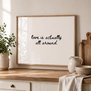 May include: A framed print with the words "love is actually all around" in black script. The frame is light brown, and the background is off-white. The print is displayed on a wooden countertop with kitchen accessories.