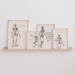 May include: A collection of framed art prints featuring black and white skeleton illustrations. Each print has a different pose and text, including "Life, man," "Dancing through life," and "The horrors persist, but so do I."