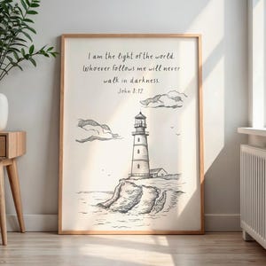 May include: A framed print featuring a black and white illustration of a lighthouse on a cliff, with text from John 8:12. The artwork is in a light wooden frame, displayed against a white wall, and includes a small house and clouds.