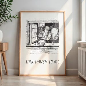 May include: A framed black and white print featuring a woman in a window, with the text "TALK DARCY TO ME" in a handwritten font. The artwork is in a light wood frame and is displayed on a wooden floor.
