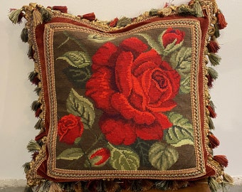 May include: A red velvet pillow with a floral design featuring a large red rose and green leaves. The pillow has a gold and red fringe trim.