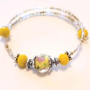 May include: A silver-toned wrap bracelet with yellow accents and a central bead featuring a pink rose design. The bracelet has multiple strands of small beads and yellow pom-pom-like beads. The central bead is clear with a yellow background.