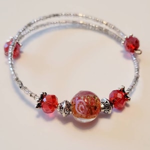 May include: A silver-toned bracelet with two strands of tiny beads. The bracelet features red faceted beads and a central glass bead with a swirling design. Decorative silver-colored metal accents are also present.