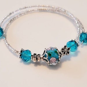 May include: A delicate silver-toned bracelet with turquoise and floral beads. The bracelet features a central, round bead with a blue background and pink floral design, complemented by faceted turquoise beads and small flower accents.