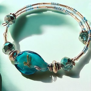 May include: A handmade bracelet with a large, turquoise glass bead shaped like a teardrop, featuring a gold dragonfly. The bracelet has faceted teal beads, silver accents, and a coiled wire design. A unique piece of jewellery.