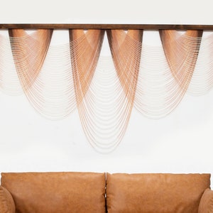 May include: A wall hanging made of woven strands of yarn in shades of peach and white. The strands are arranged in a cascading pattern, creating a wave-like effect. The hanging is mounted on a wooden frame.